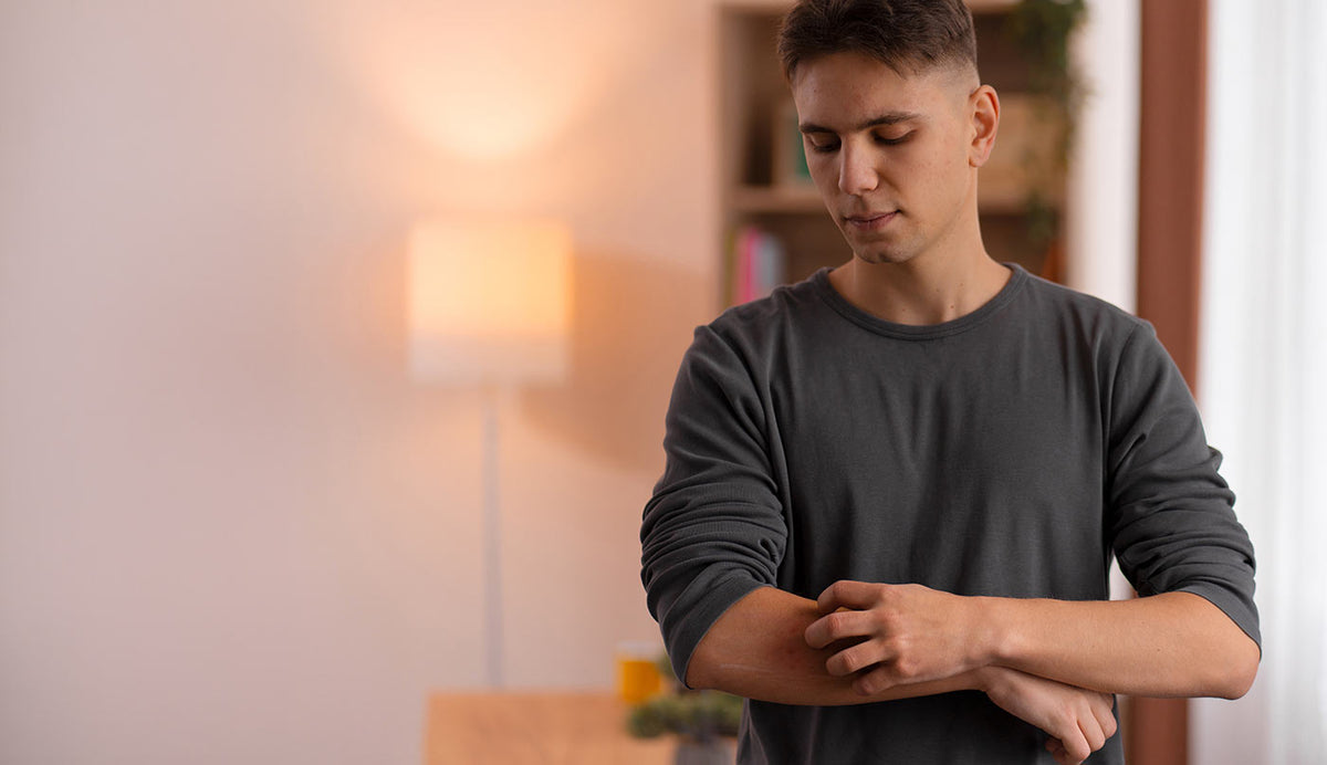 A man in a dark gray long-sleeve shirt standing indoors and rubbing his arm, with a softly lit room and blurred lamp in the background.