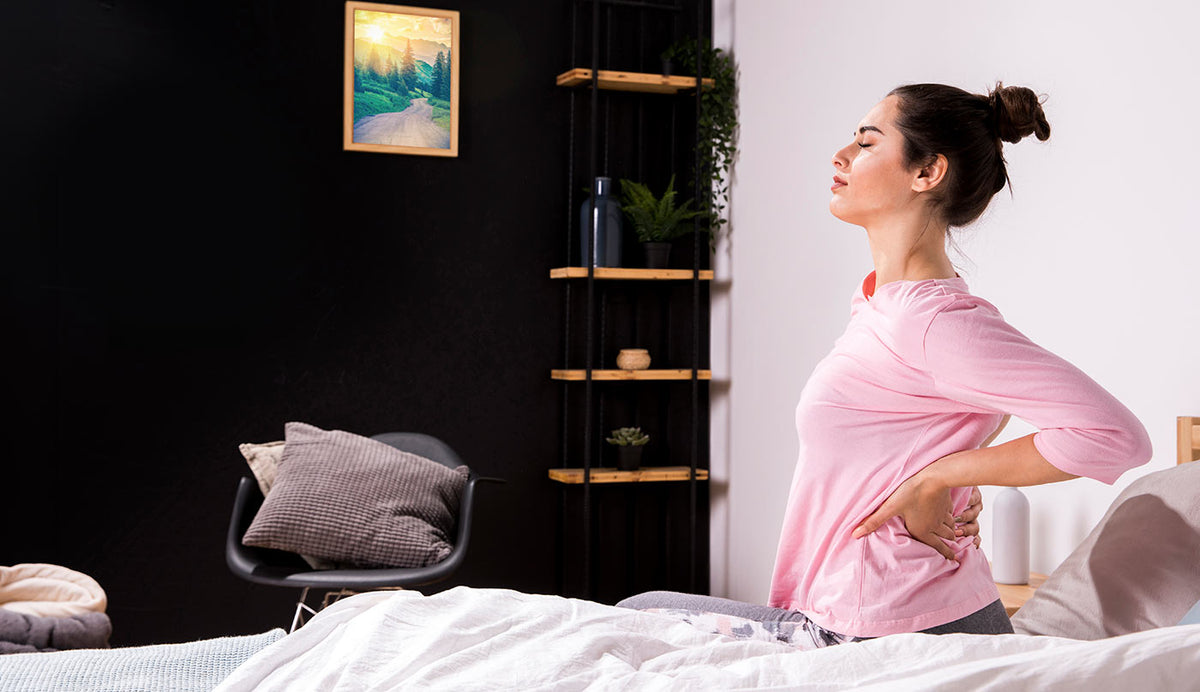 A woman in a pink long-sleeve shirt stands on a bed in a bedroom, holding her lower back in a stretching pose. A black chair with a gray pillow is on the left, and a black shelving unit with wooden shelves is on the right against a white wall.
