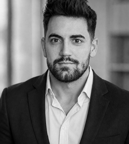 A black and white portrait of Dr Ben with a beard and styled hair, wearing a dark suit jacket over a light dress shirt, shown from the chest up against a softly blurred indoor background.