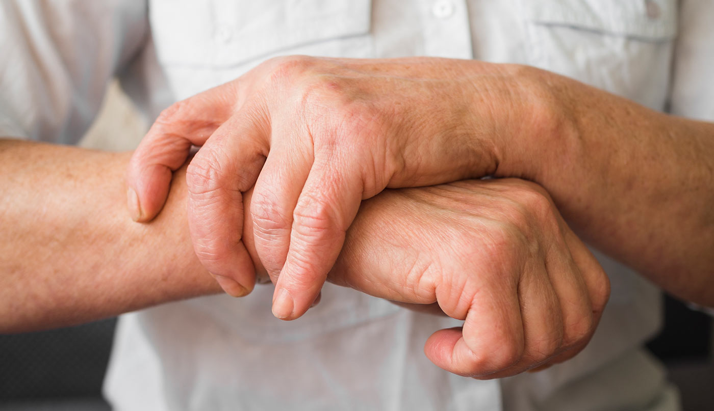 Close-up of an older person's hands and forearms, with one hand holding and pressing on the other wrist area. The person is wearing a white button-up shirt.