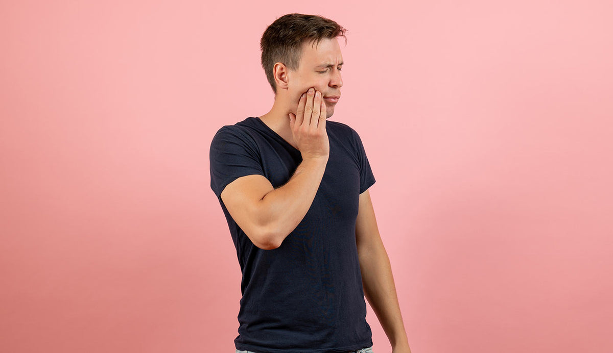 A man in a dark navy blue t-shirt stands against a solid pink background, holding his hand to his cheek in a gesture of pain or discomfort.
