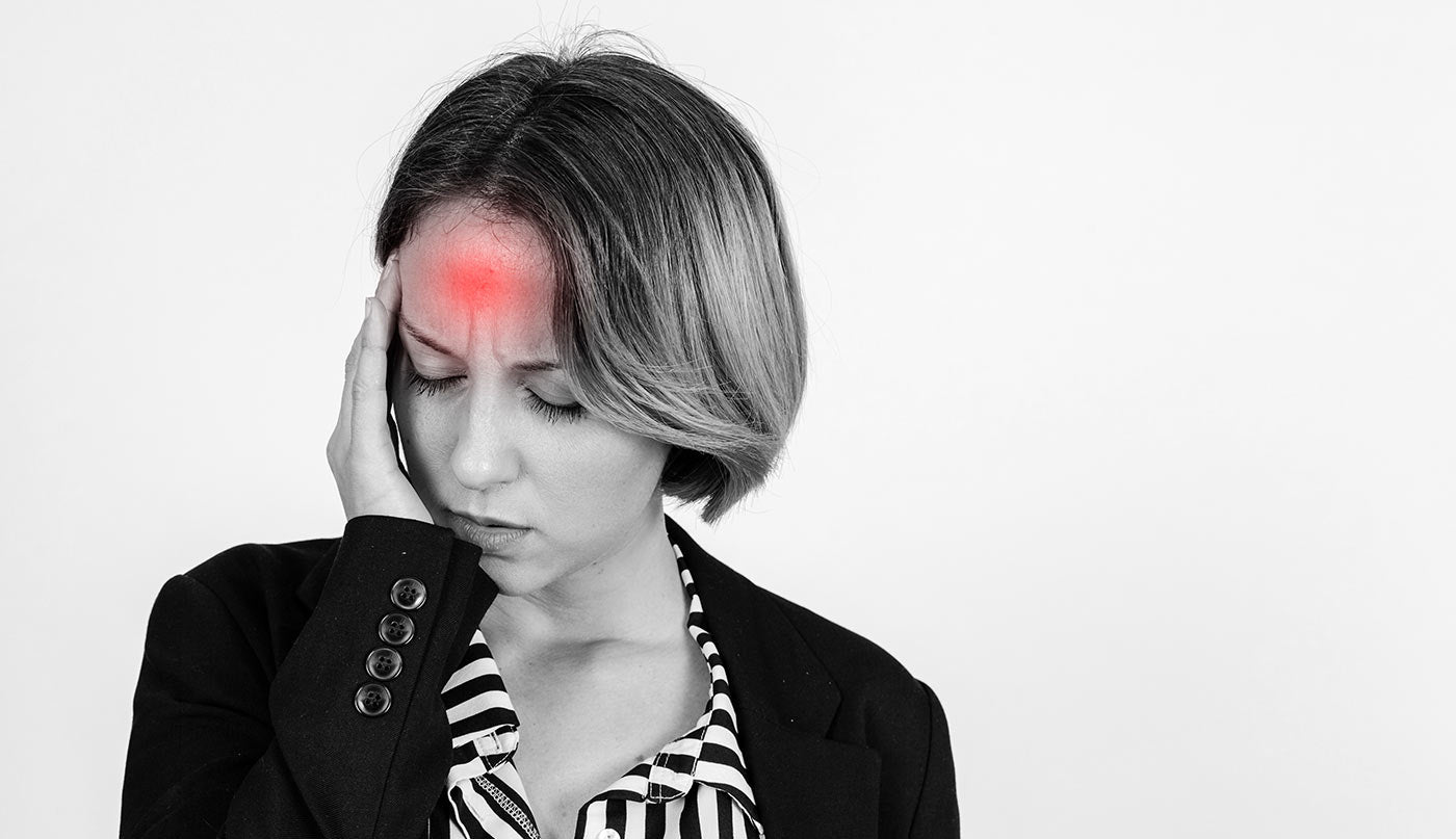 A woman with short gray hair, wearing a black blazer and a black-and-white striped shirt, holding her head with her hand. A red glow highlights her forehead, set against a plain white background.