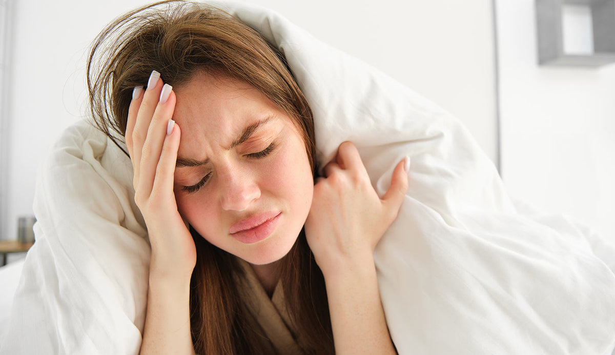 A woman lying in bed under white covers, holding her head with one hand and shielding her ear with the other, appearing distressed or uncomfortable in a softly lit bedroom.