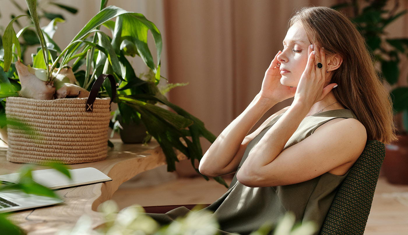 A woman sits in a chair in a home office environment, holding her temples as if experiencing stress or headaches. She is wearing a sleeveless olive green top.