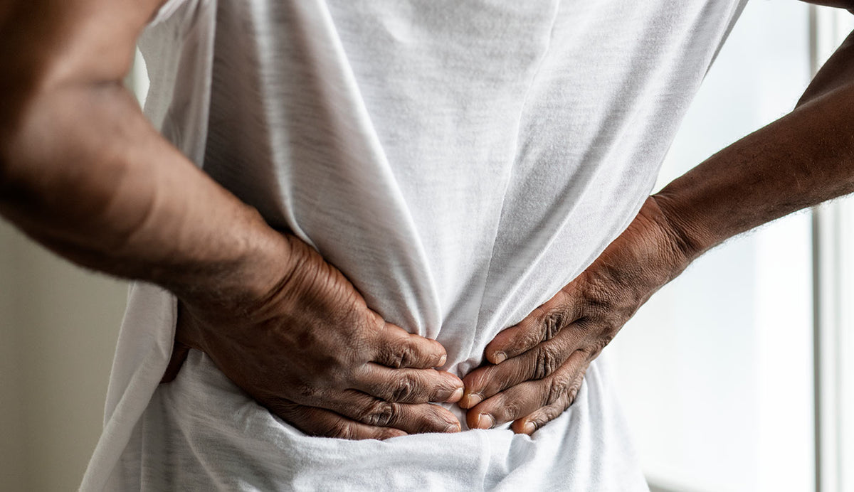 A close-up shot of a person in a white t-shirt holding their lower back in pain, with their hands on their hips against a softly lit indoor background.
