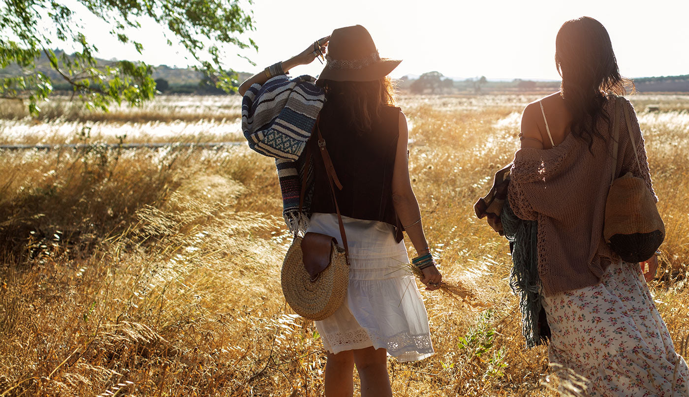 Two women walking through a field with one carrying a bag and the other a hat.